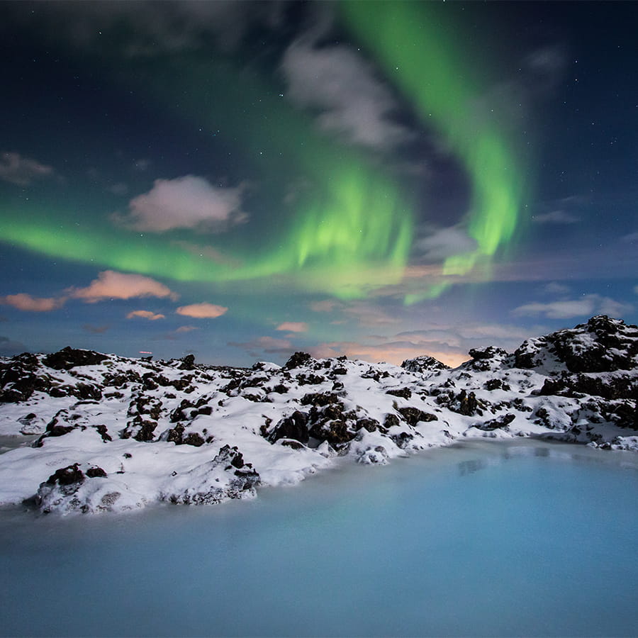 Green and blue northern lights above the Blue Lagoon in Iceland with geothermal steam rising from warm waters in a snowy winter landscape