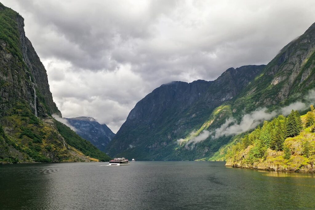 Cruise boat gliding through the narrow waters of Nærøyfjord in Norway surrounded by towering cliffs and deep green reflections in the fjord