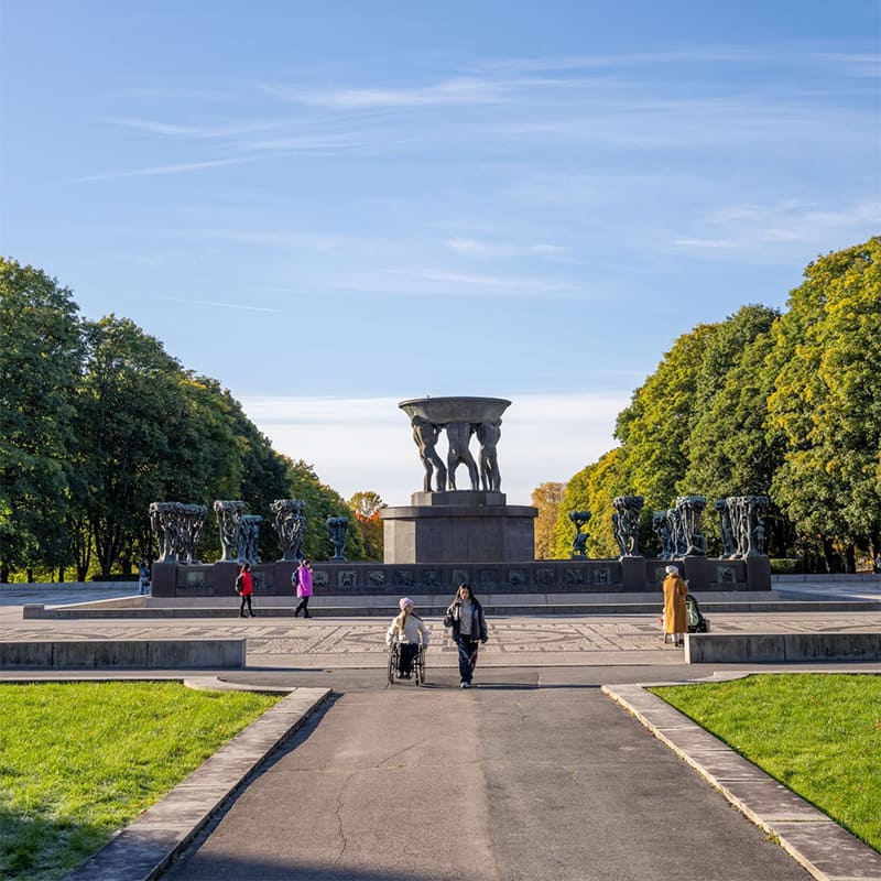Visitors walking among expressive stone sculptures in the Vigeland sculpture park in Oslo surrounded by green lawns and open sky