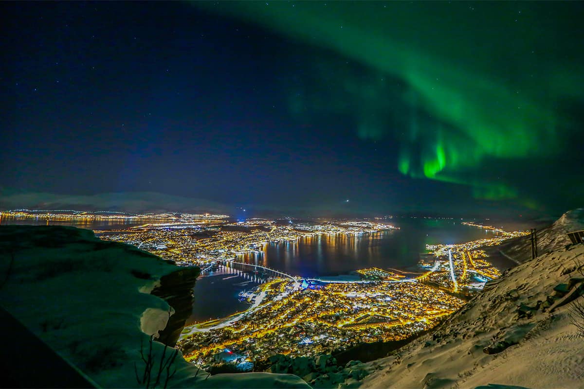 The northern lights shimmer above snowy Tromsø in winter, casting a magical glow over the Arctic cityscape and surrounding mountains